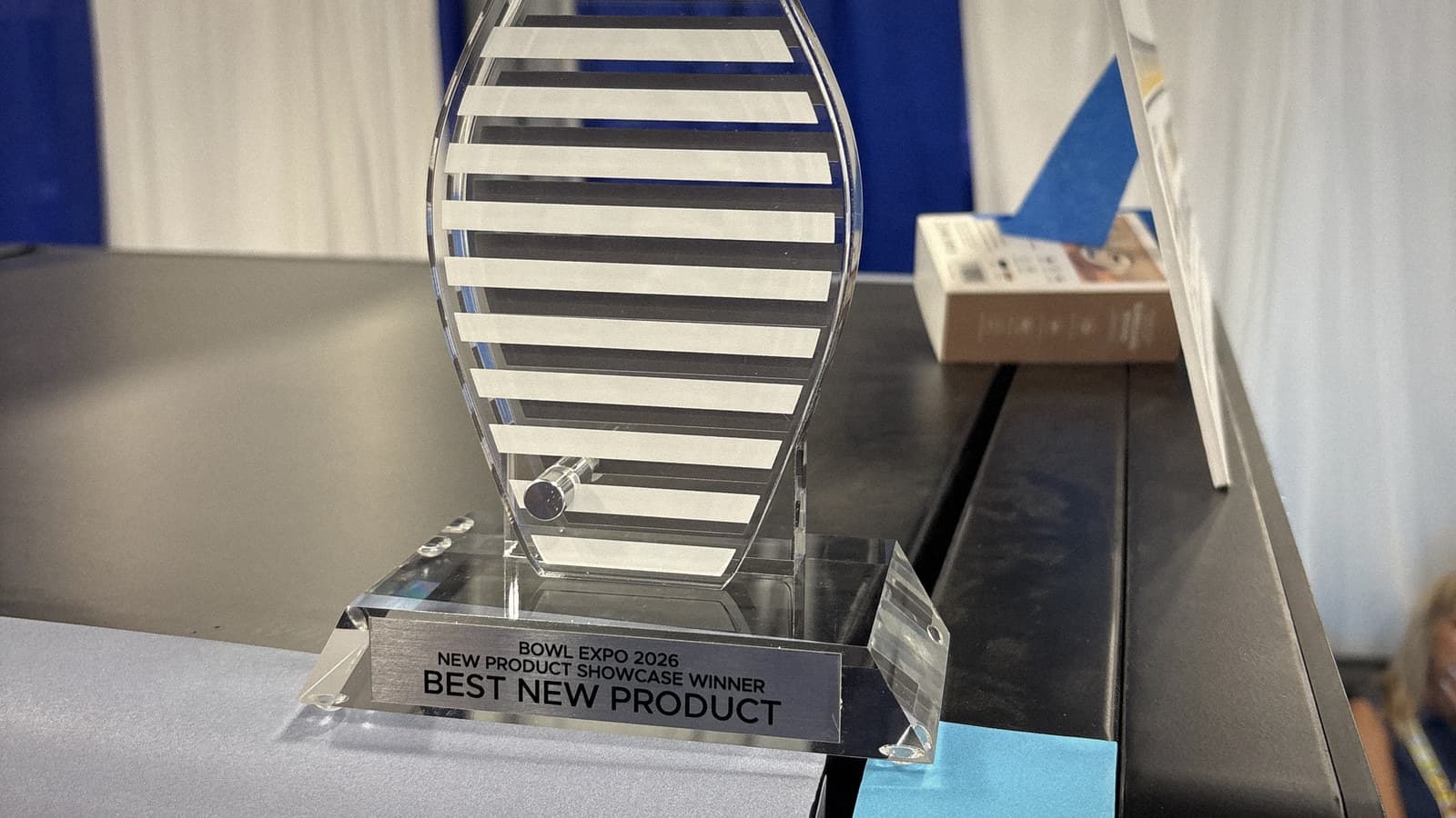 The Bowl Expo 2026 Best New Product trophy, a clear acrylic bowling pin, standing on the Custom Case Kiosk at the show in Nashville.