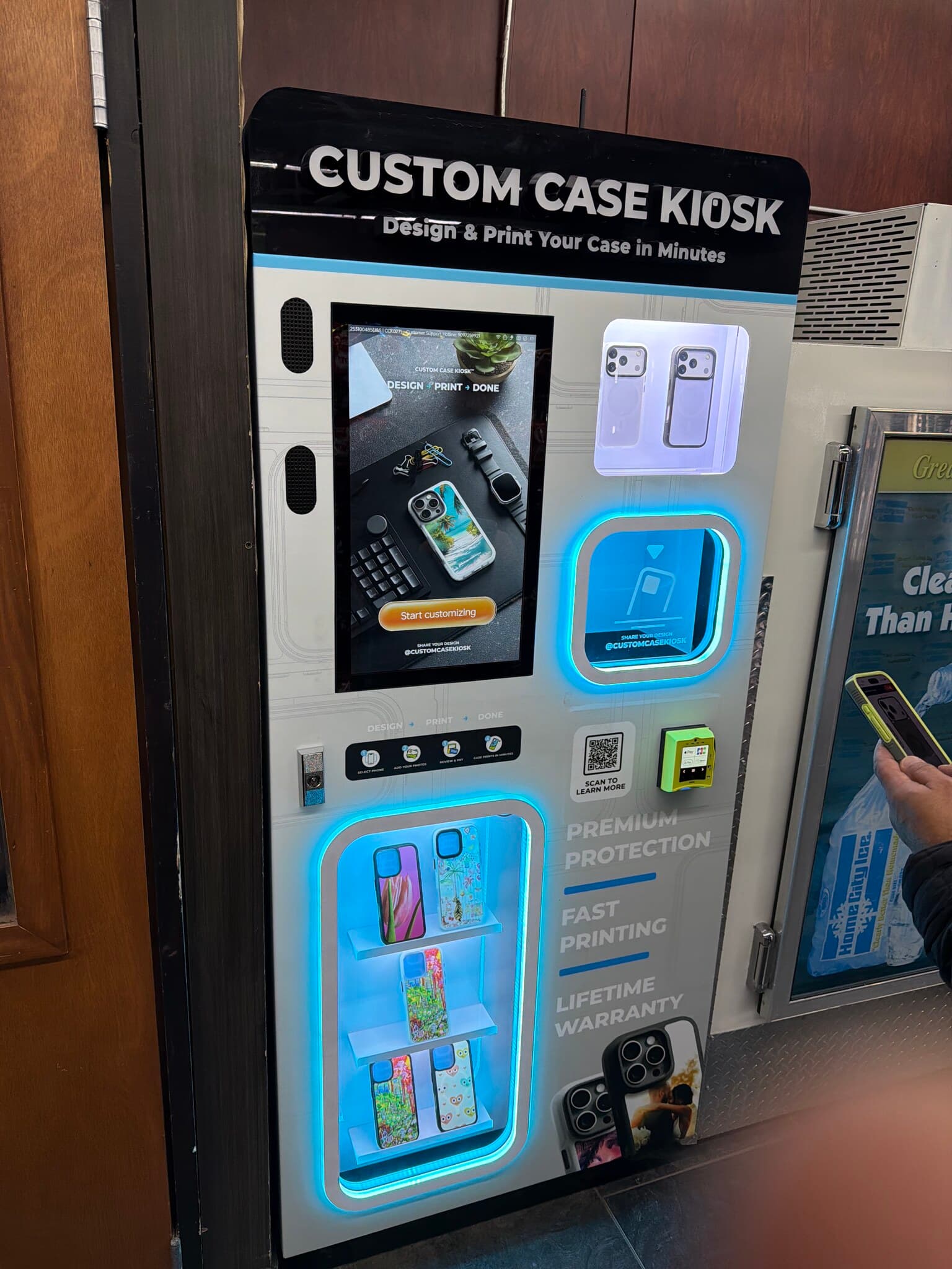 Custom Case Kiosk at 10 E McNichols Rd in Highland Park, Michigan — the kiosk lit up against the store's wood-panelled wall, printed cases on the backlit shelves