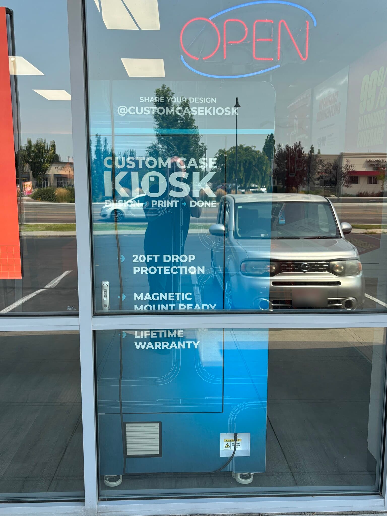 The Custom Case Kiosk seen from the sidewalk through the Boost Mobile storefront window in Nampa, Idaho, below the store's neon OPEN sign