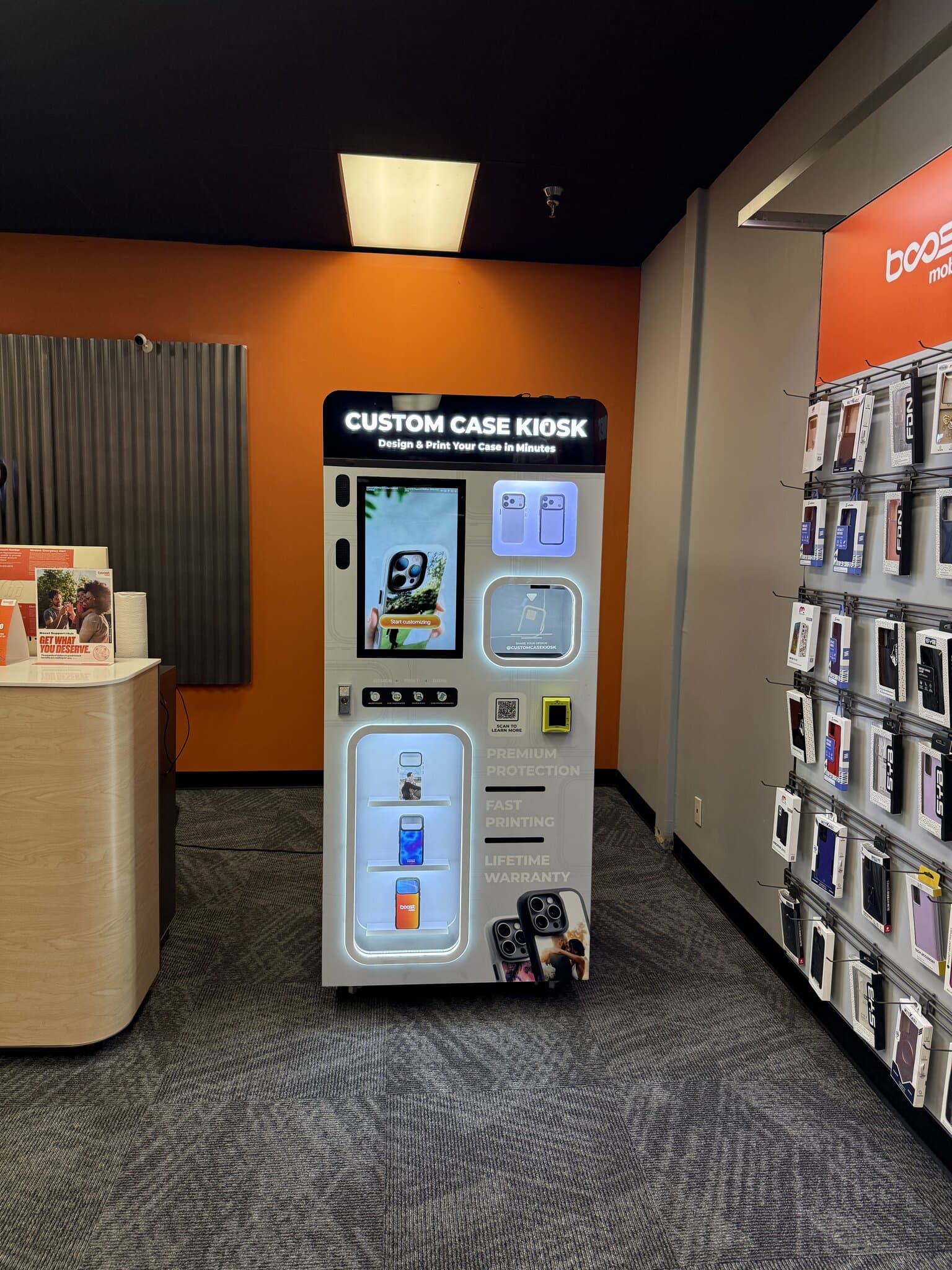 Custom Case Kiosk against the orange accent wall inside the Boost Mobile store in Phoenix, AZ — lit design touchscreen, case pickup window, and printed-case display shelves