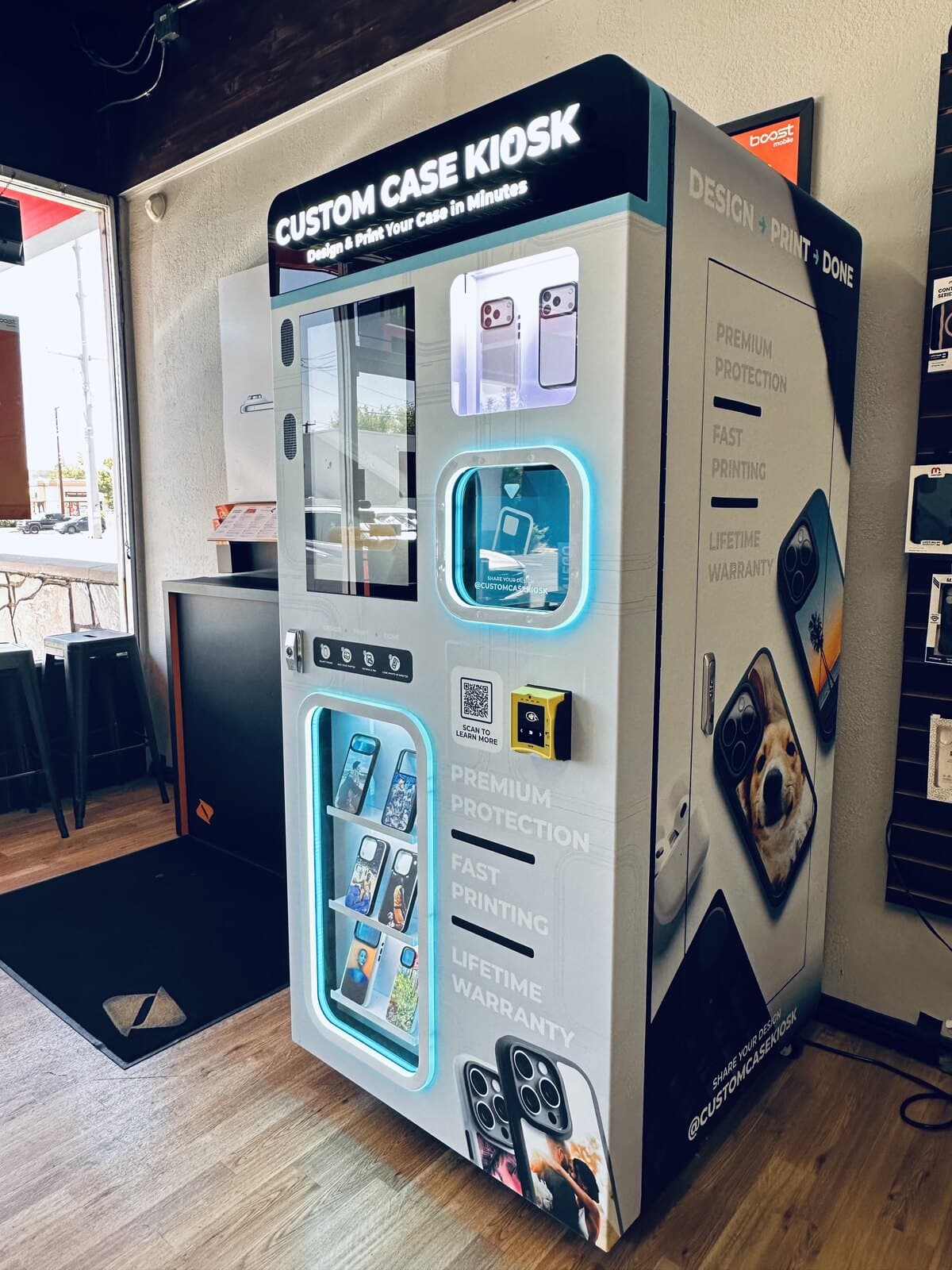 The Custom Case Kiosk at Boost Mobile in Salt Lake City seen from the side, its lit marquee above a backlit shelf of printed cases, next to the Design, Print, Done panel